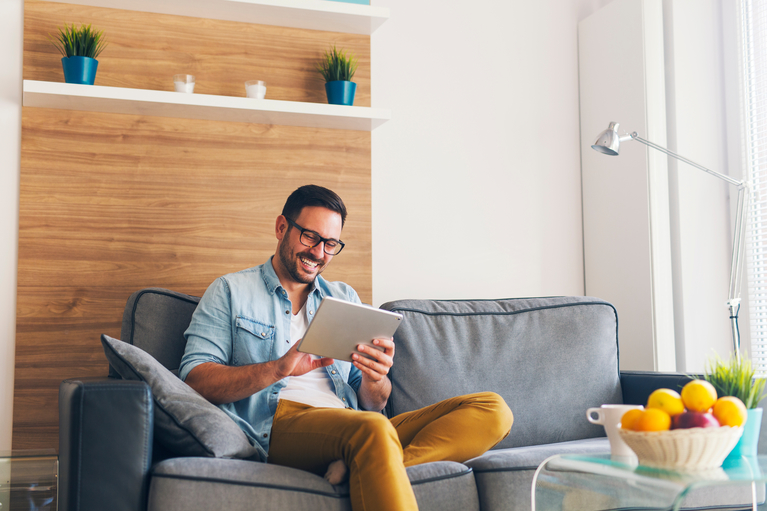 Ein Mann sitzt glücklich auf seiner Couch und benutzt sein Tablet.